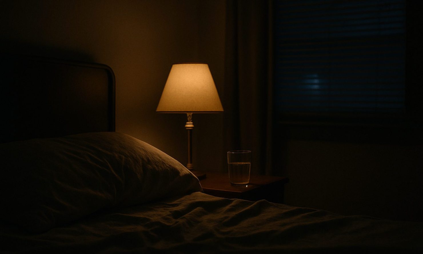A dark bedroom, bedside lamp on over a rumpled pillow and a glass of water on the nightstand